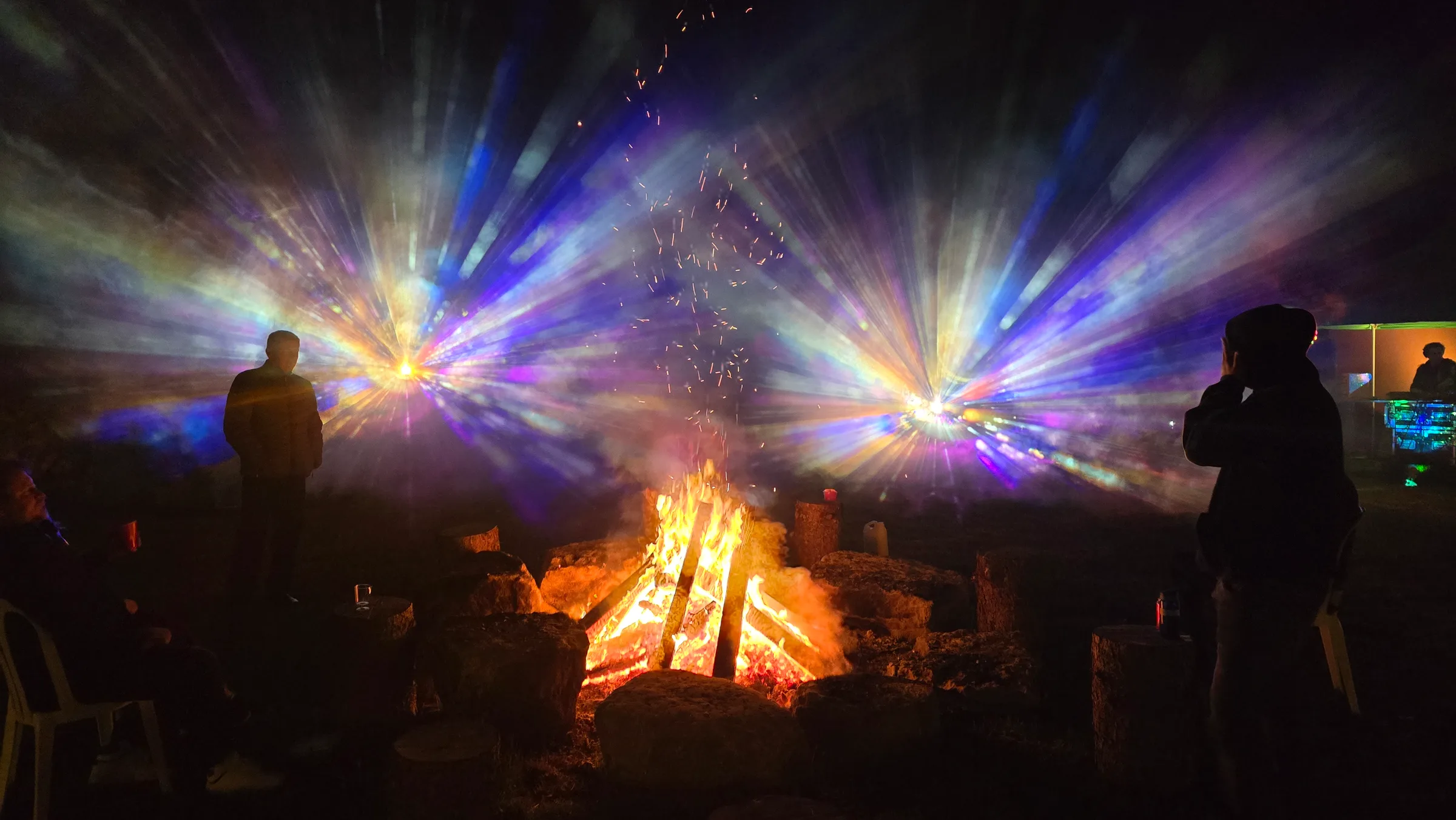 Silhouetted people gather around a bonfire at night as twin starburst beams of blue, purple, and gold light pierce smoky haze overhead.