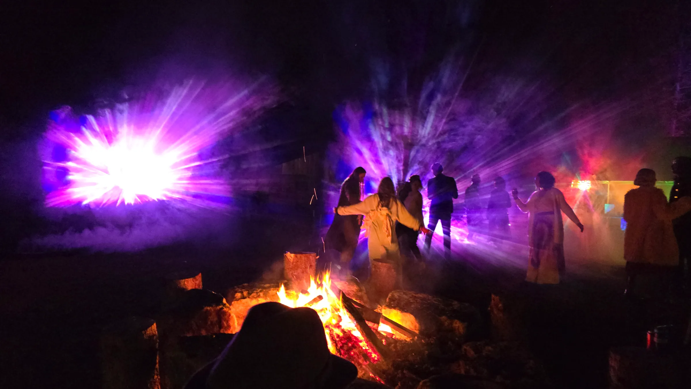 A blurred silhouette in a hat watches a bonfire while a crowd dances nearby, lit by an intense white-purple light burst and fanning purple beams.