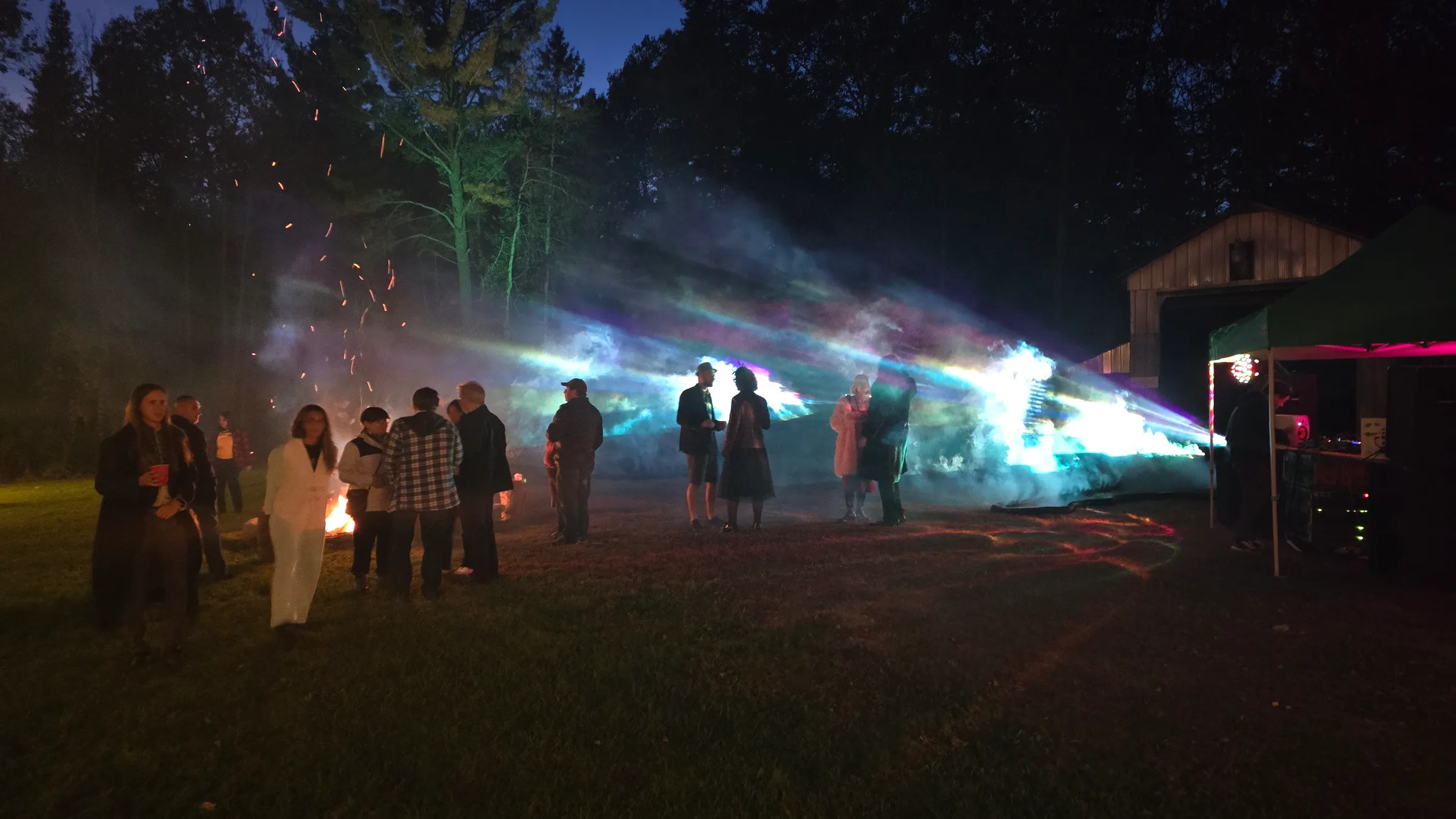 A row of onlookers stands at night as parallel beams of blue, magenta, and white light cut horizontally through fog in front of a wooden barn.
