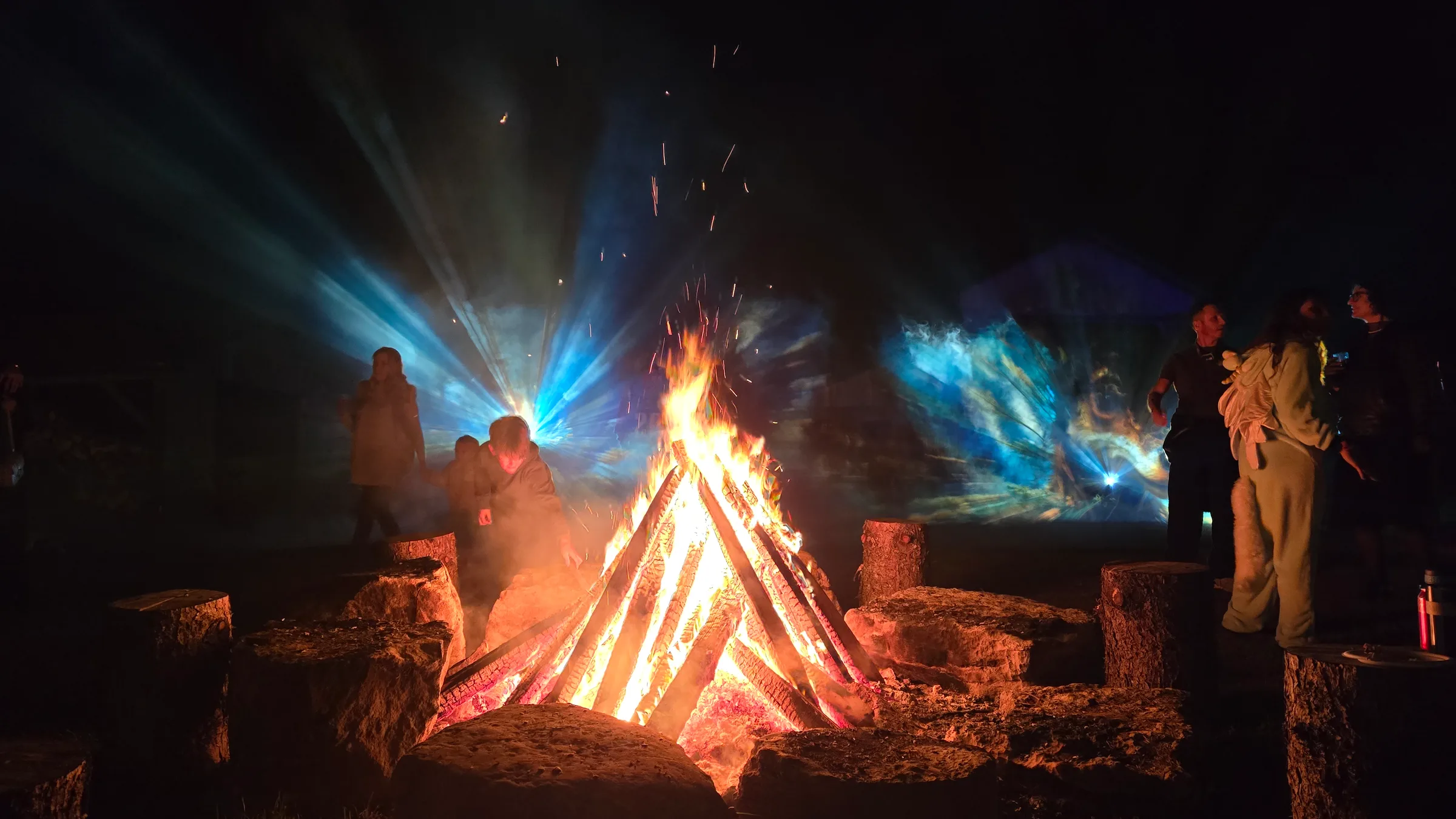 Silhouetted figures near a tall bonfire include someone in a fuzzy costume, with blue light rays and a glowing teal cloud filling the night air.