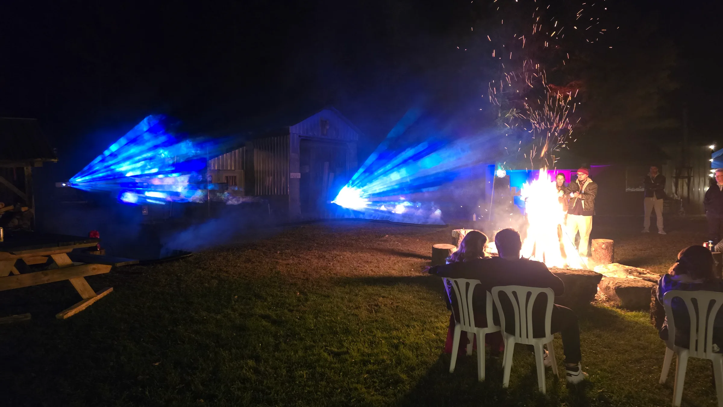 Seated onlookers in patio chairs watch a bonfire and two diagonal blue light beams piercing the fog in front of a dark barn at night.