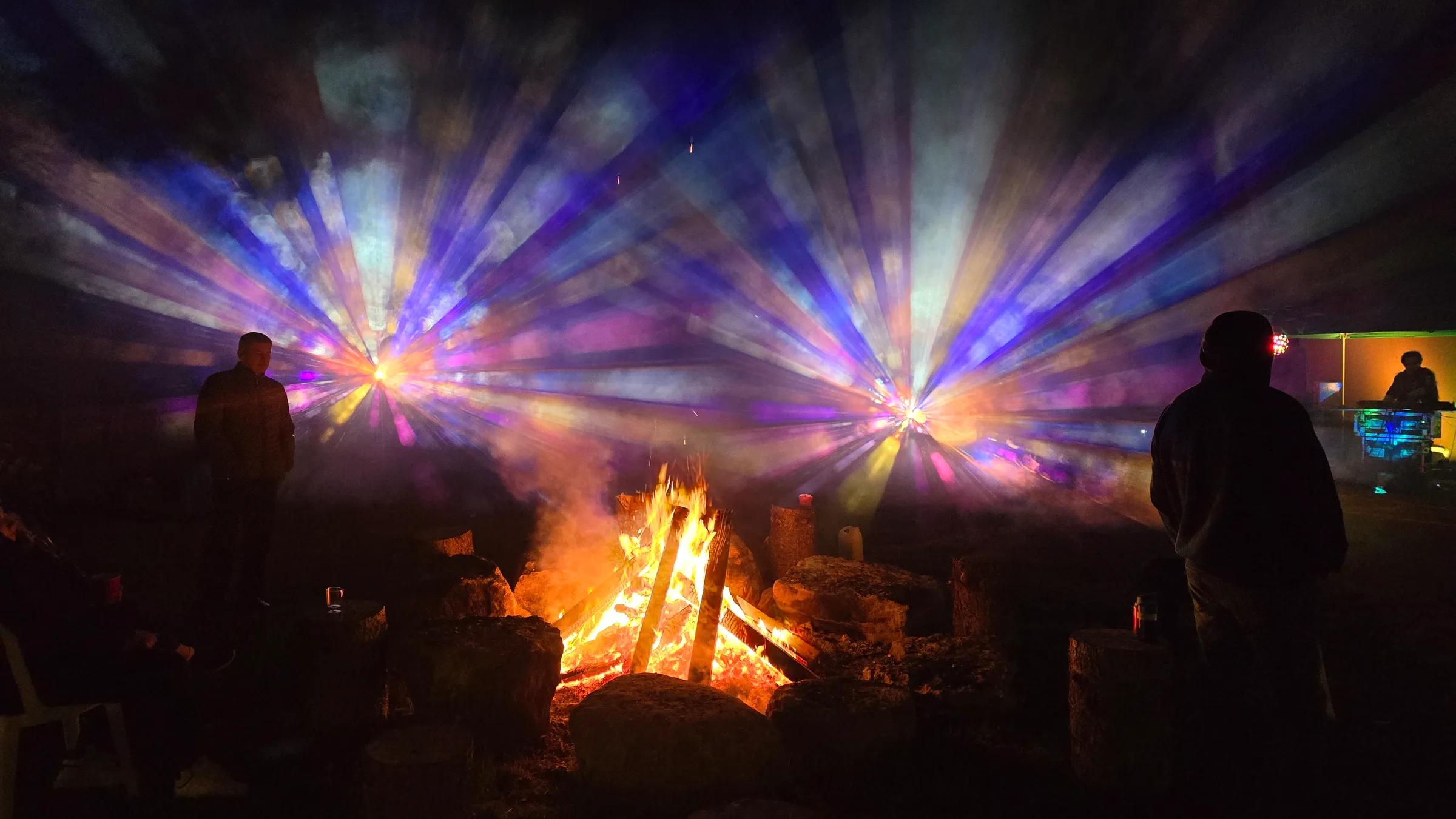 Two silhouetted figures flank a bonfire as symmetrical bursts of warm and blue light radiate through smoky haze, with lit equipment glowing nearby.